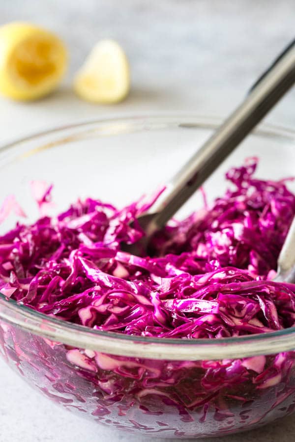 A bowl of red cabbage slaw with kitchen tongs sticking out. 