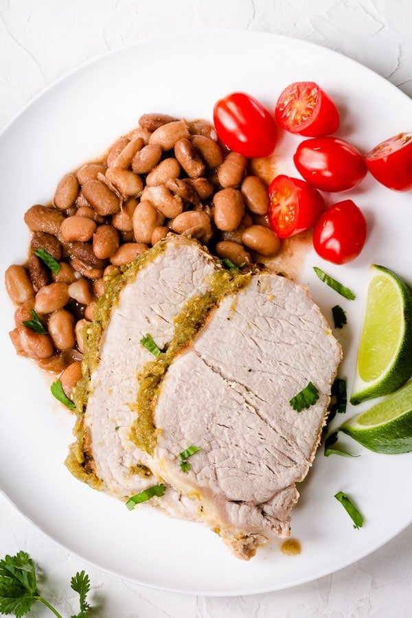 Instant Pot salsa verde pork loin slices on a plate with pinto beans, tomatoes, and lime wedges. 