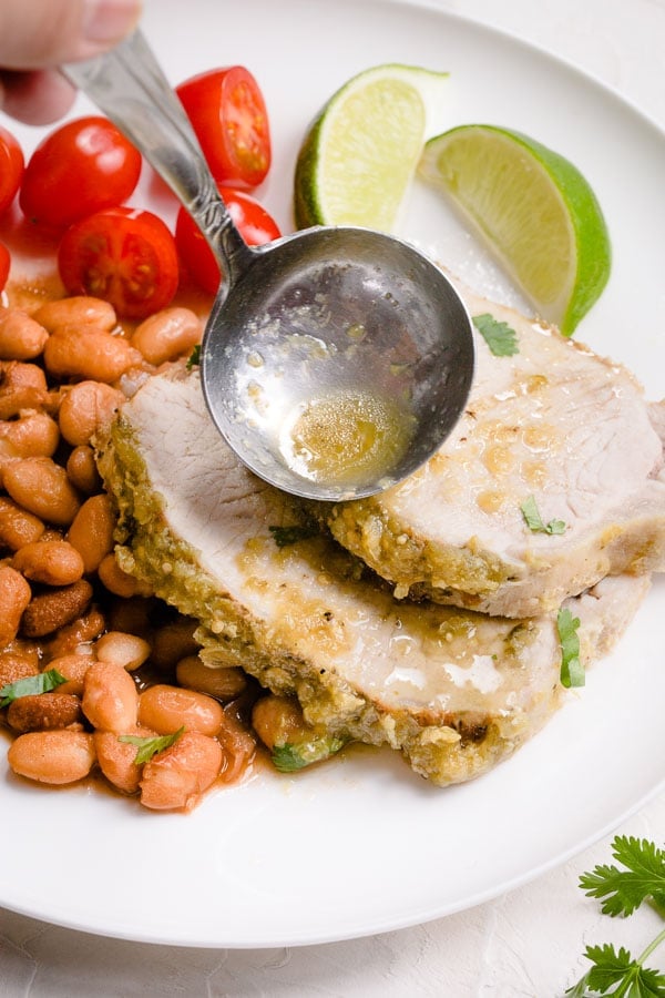 Salsa verde juice being poured over a pork loin roast on a plate with pinto beans, tomatoes, and lime wedges. 