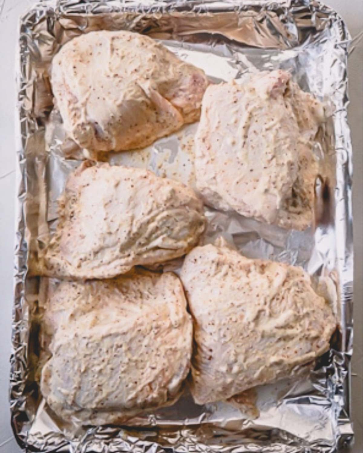 Five chicken thighs coated with mayo mixture are layed out on a foil lined baking sheet.