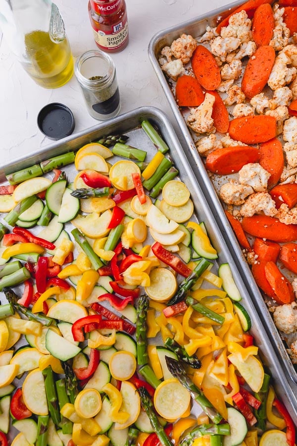 Two baking sheets of roasted vegetables side by side with spices and oil at the top.