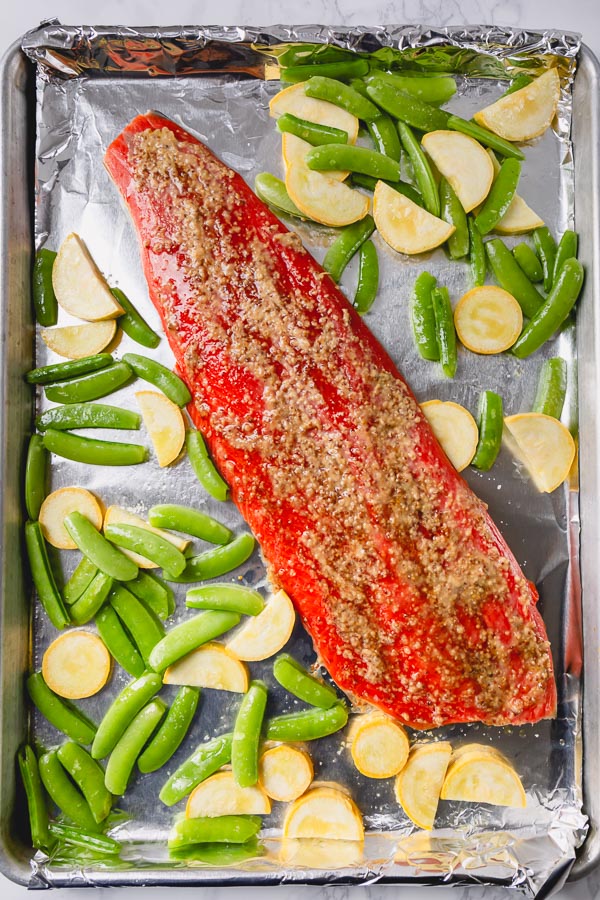 A sheet pan lined with foil topped with raw salmon, sugar snap peas, and squash. 