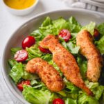 3 crispy chicken fingers over a bed of lettuce and cherry tomatoes.