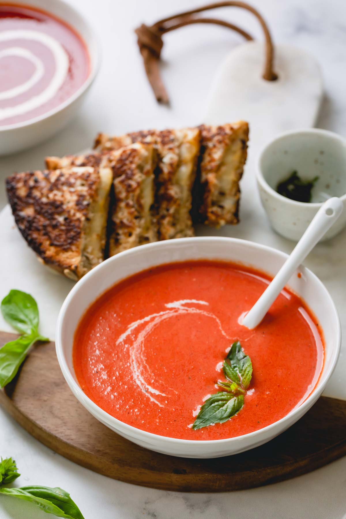 A bowl of smooth roasted red bell pepper soup garnished with cream and basil leaves and side of grilled cheese.