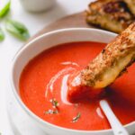 A bowl of roasted red pepper soup with grilled cheese dipped in it.