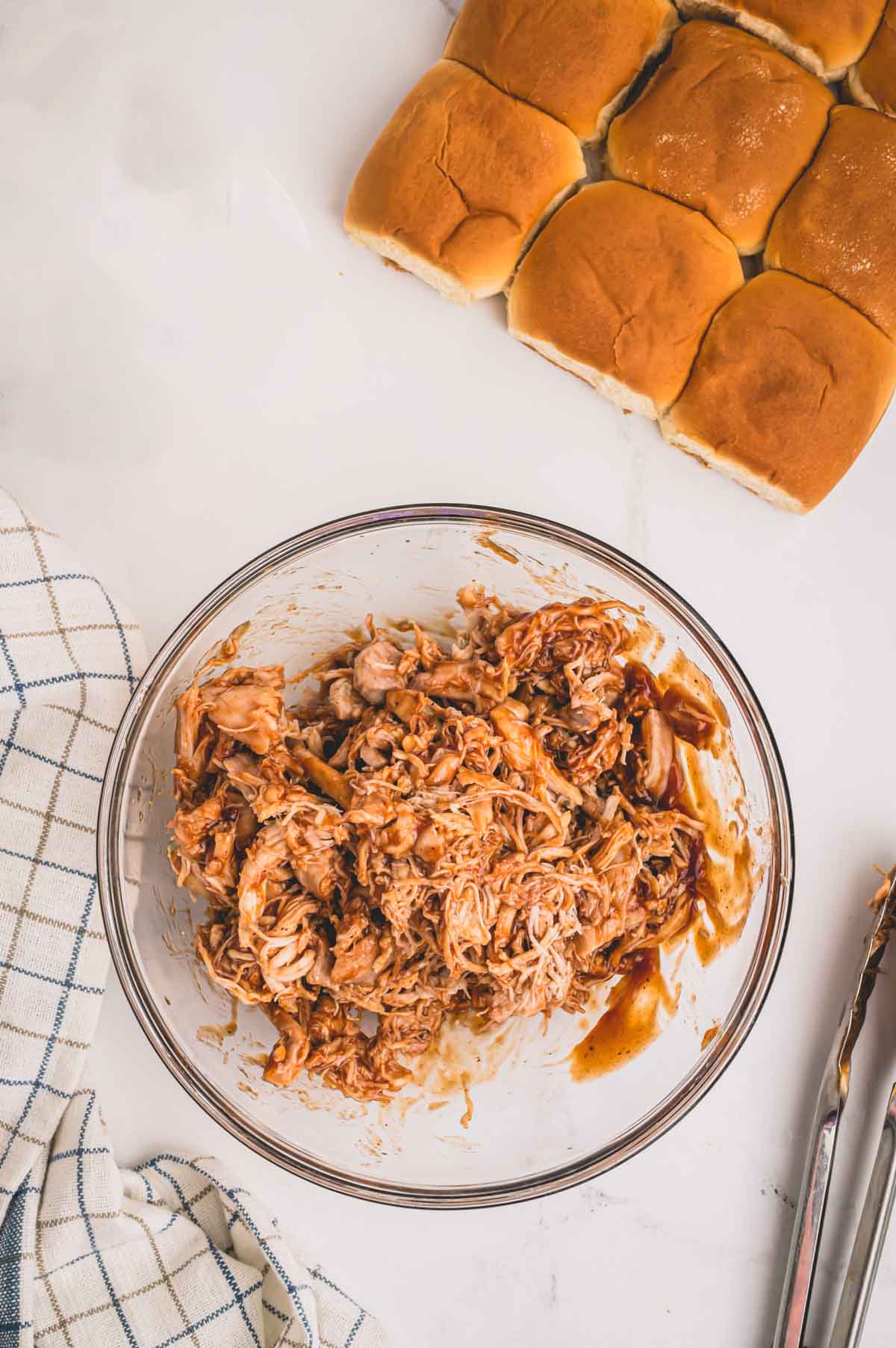 Shredded chicken mixed with bbq sauce in a glass bowl and slider rolls on the side.