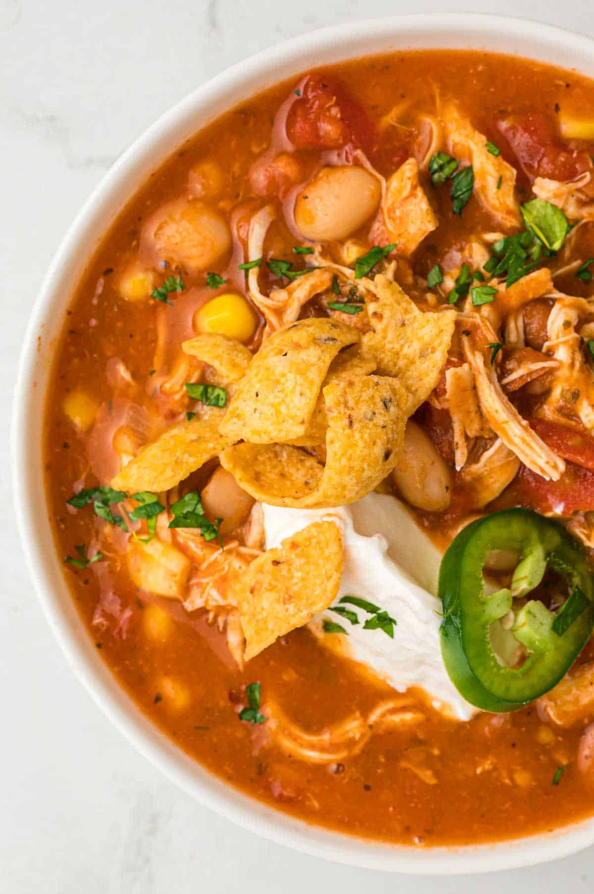 A bowl of chicken chili topped with frito chips, sour cream, jalapeno slice and chopped herbs.