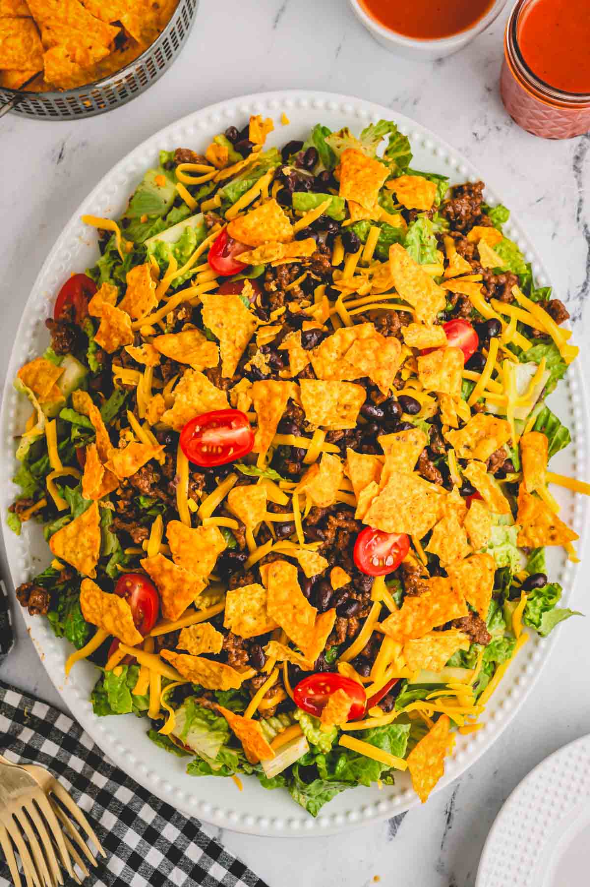 A platter of Dorito taco salad with chips, condiments, and serving utensils on the side.