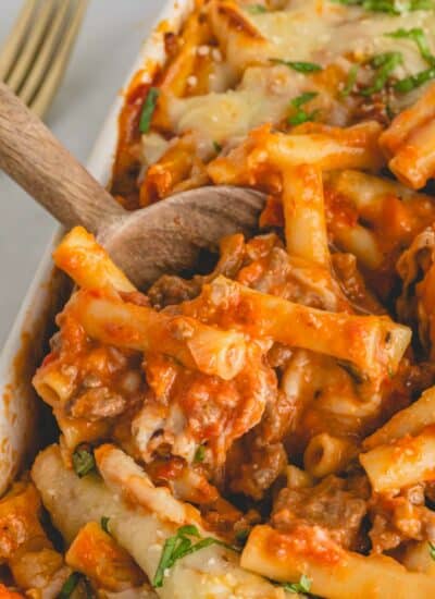 A wooden spoon dipping into a baking dish full of easy baked ziti with sausage.