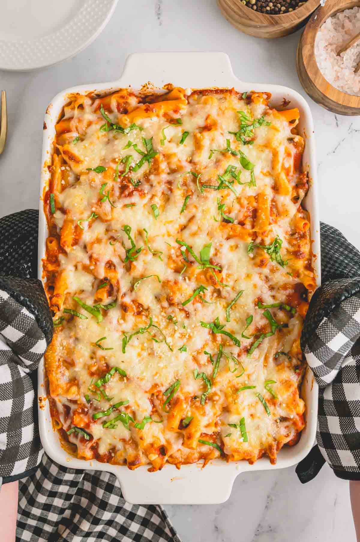 Overhead image of two holding a baking dish full of baked ziti with sausage with oven mitts.