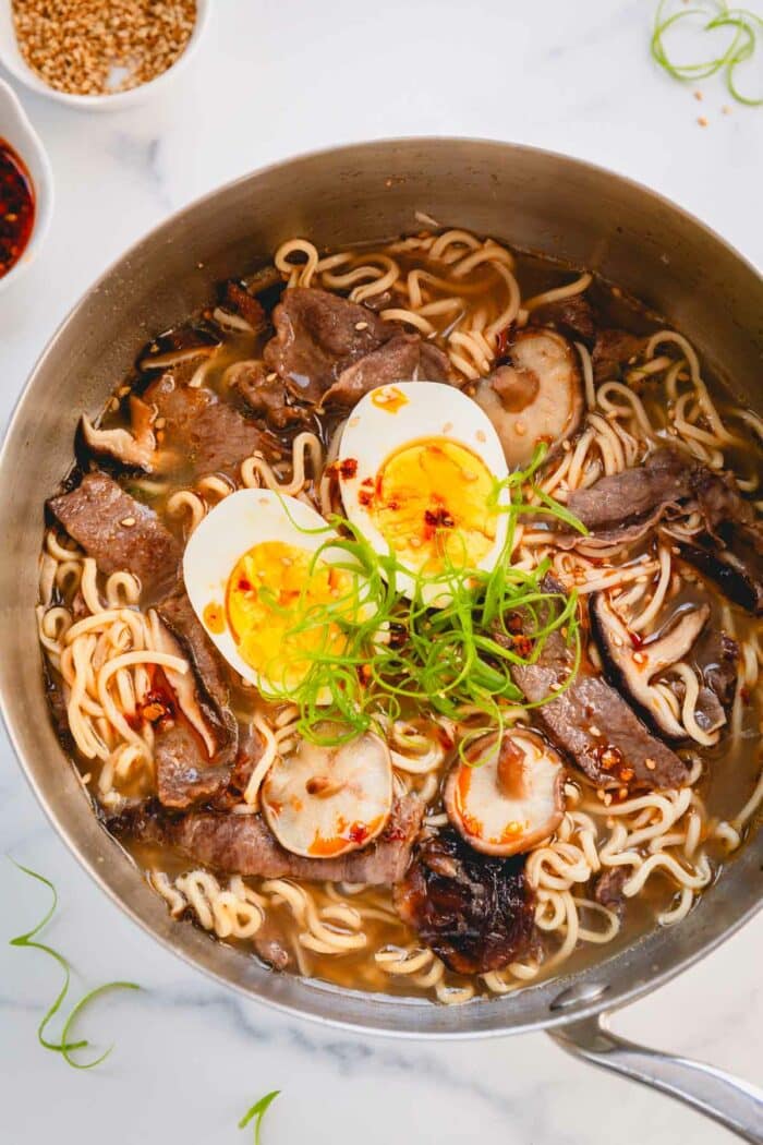 A bowl of beef ramen topped with a hard boiled egg, chopped scallions, and red pepper flakes.