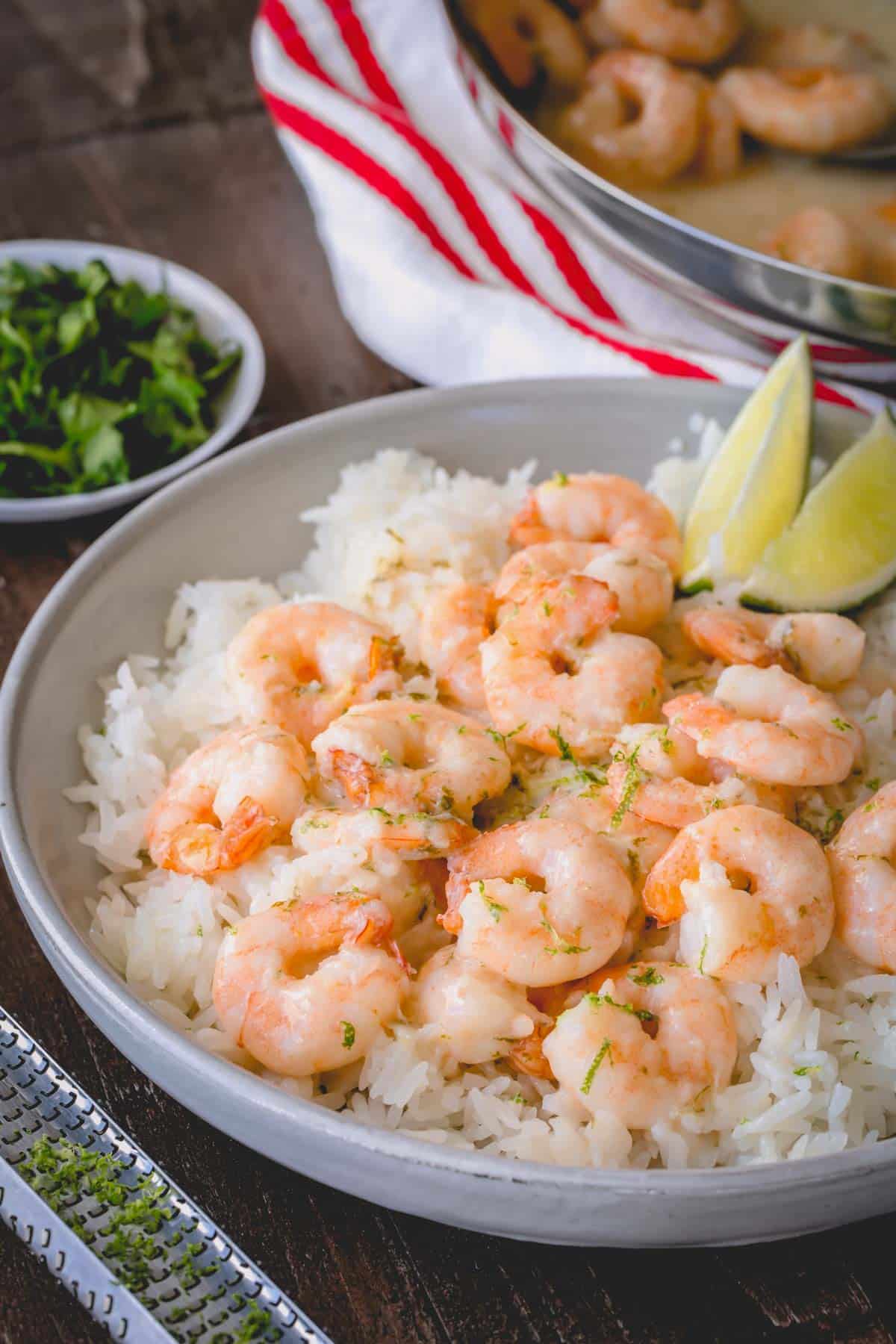 A bowl of white rice topped with creamy coconut shrimp and two lime wedges.