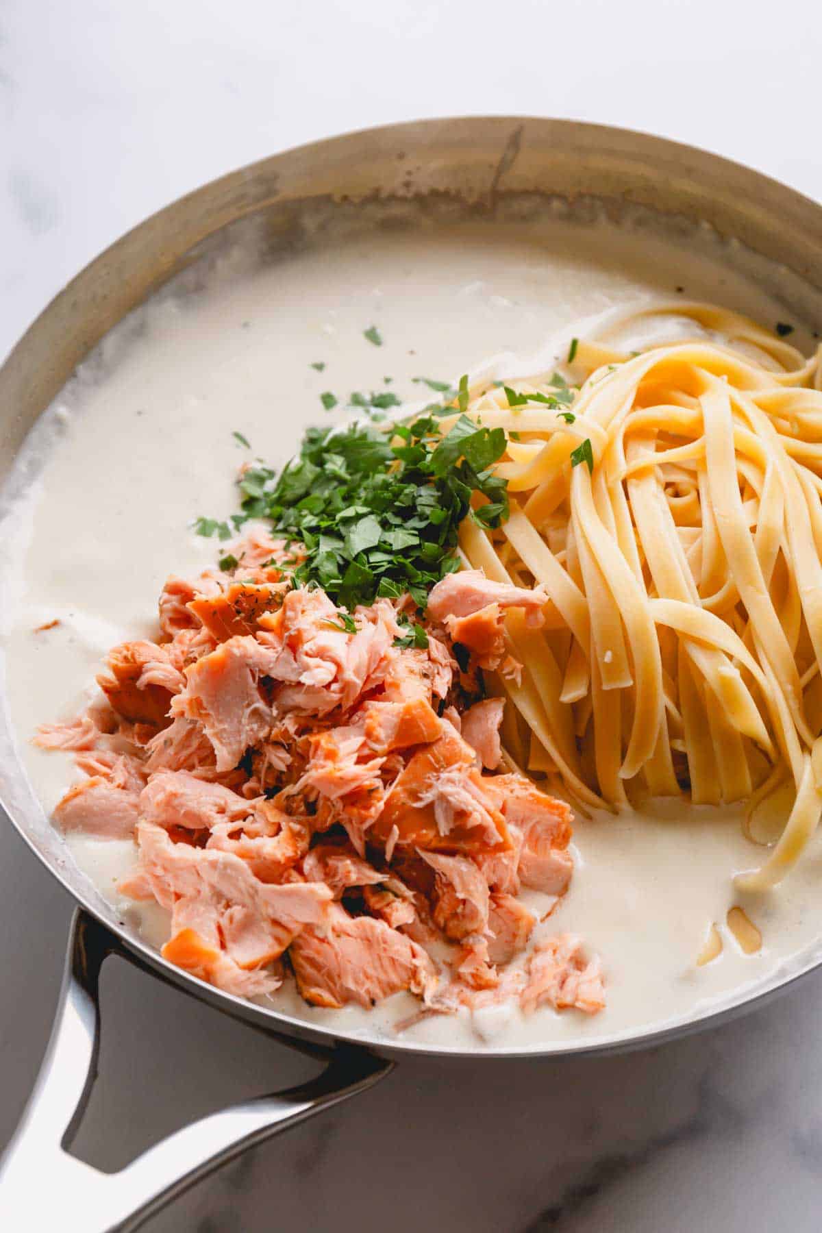 Smoked salmon, cooked fettuccine noodles, and parsley being added to alfredo sauce in a skillet.