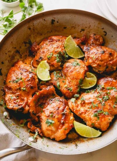 A skillet of cooked cilantro lime chicken thighs topped with lime wedges.