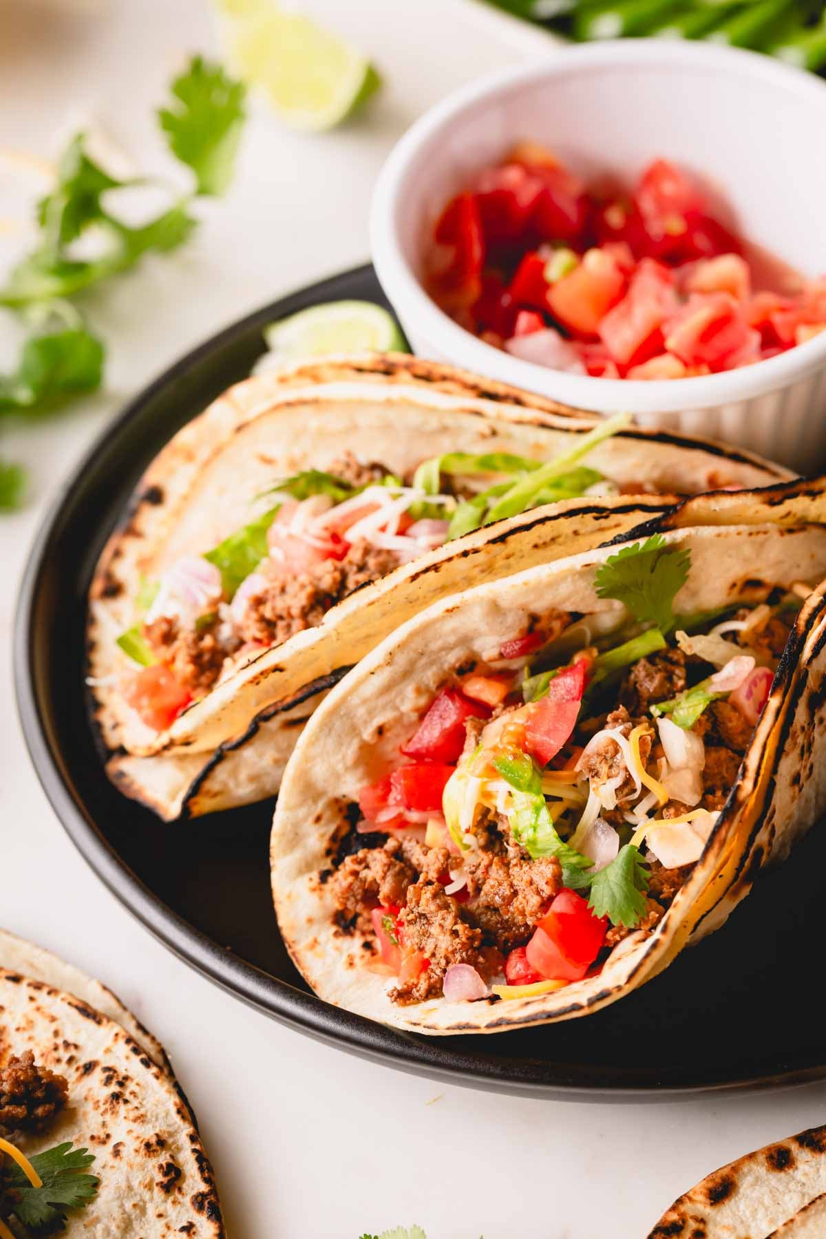 Two ground beef tacos on a plate with a small bowl of pico de gallo.
