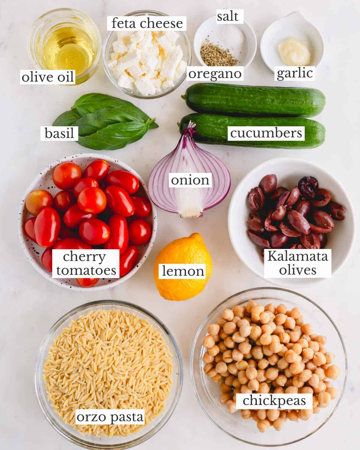 Ingredients to make a Greek orzo salad recipe. They include olive oil, feta, dried oregano, garlic, basil, cucumber, cherry tomatoes, shallot, lemon, kalamata olives, orzo, and chickpeas. 