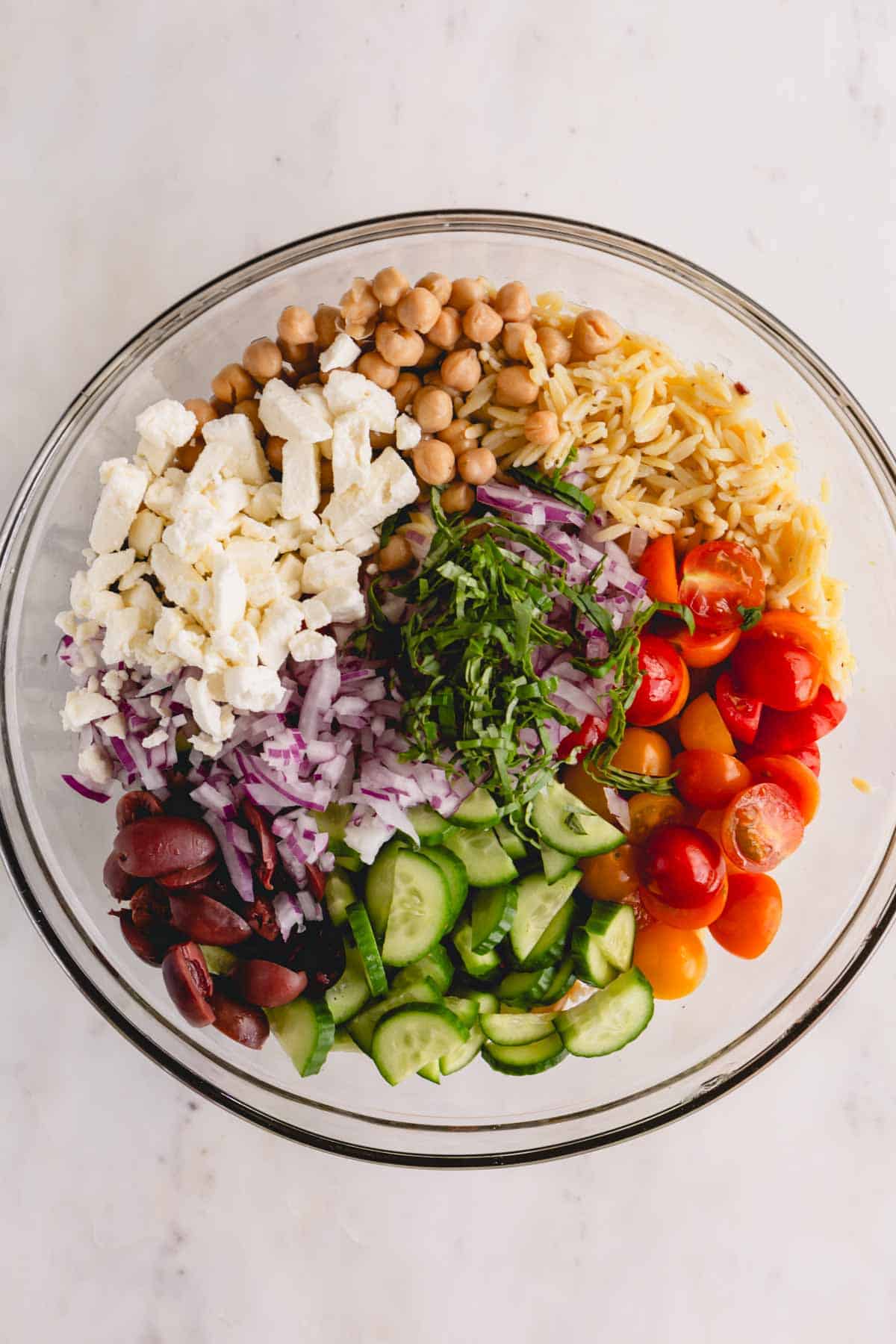 A glass bowl of cooked orzo, tomatoes, cucumbers, olives, red onion, feta, chickpeas, and herbs.