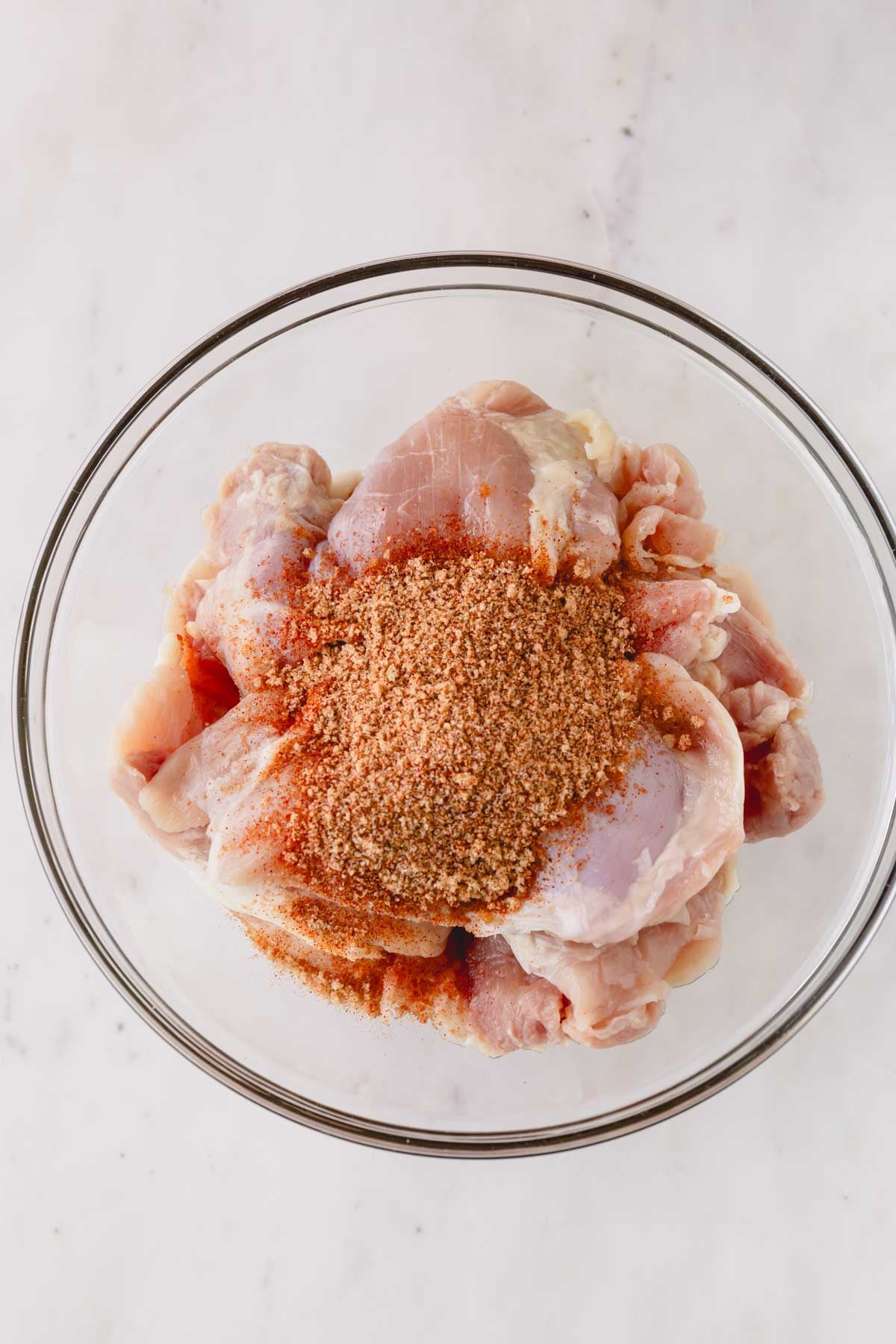 Raw chicken thighs in a bowl with a seasoning mix on top.