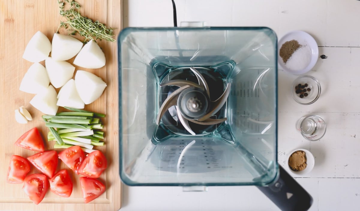Overhead image of a blender, tomatoes, green onions, white onions, herbs, and spices for jerk sauce.
