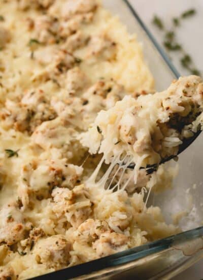A spoon lifting a scoop of cheesy chicken and rice casserole from a baking dish.