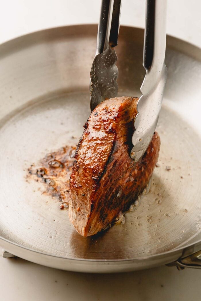 Tongs holding a seared ahi tuna steak on its side to sear the sides on a stainless steel skillet.