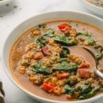 A close-up image of ham and lentil soup in a white bowl with a metal spoon. A second bowl of soup is in the background as well as a small bowl of chopped parsley.