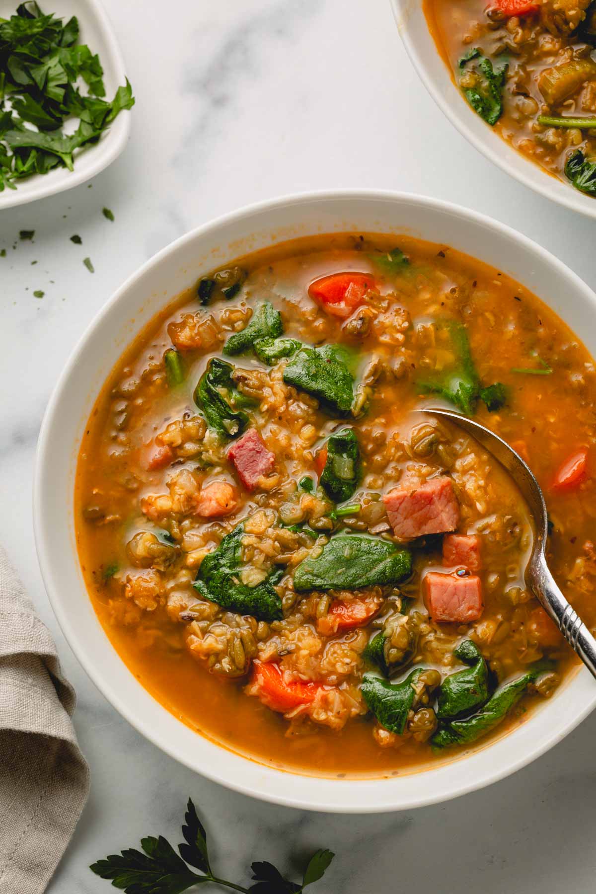 Ham and lentil soup in a white bowl with a metal spoon. A second bowl of soup in the back as well as a small bowl of chopped parsley.