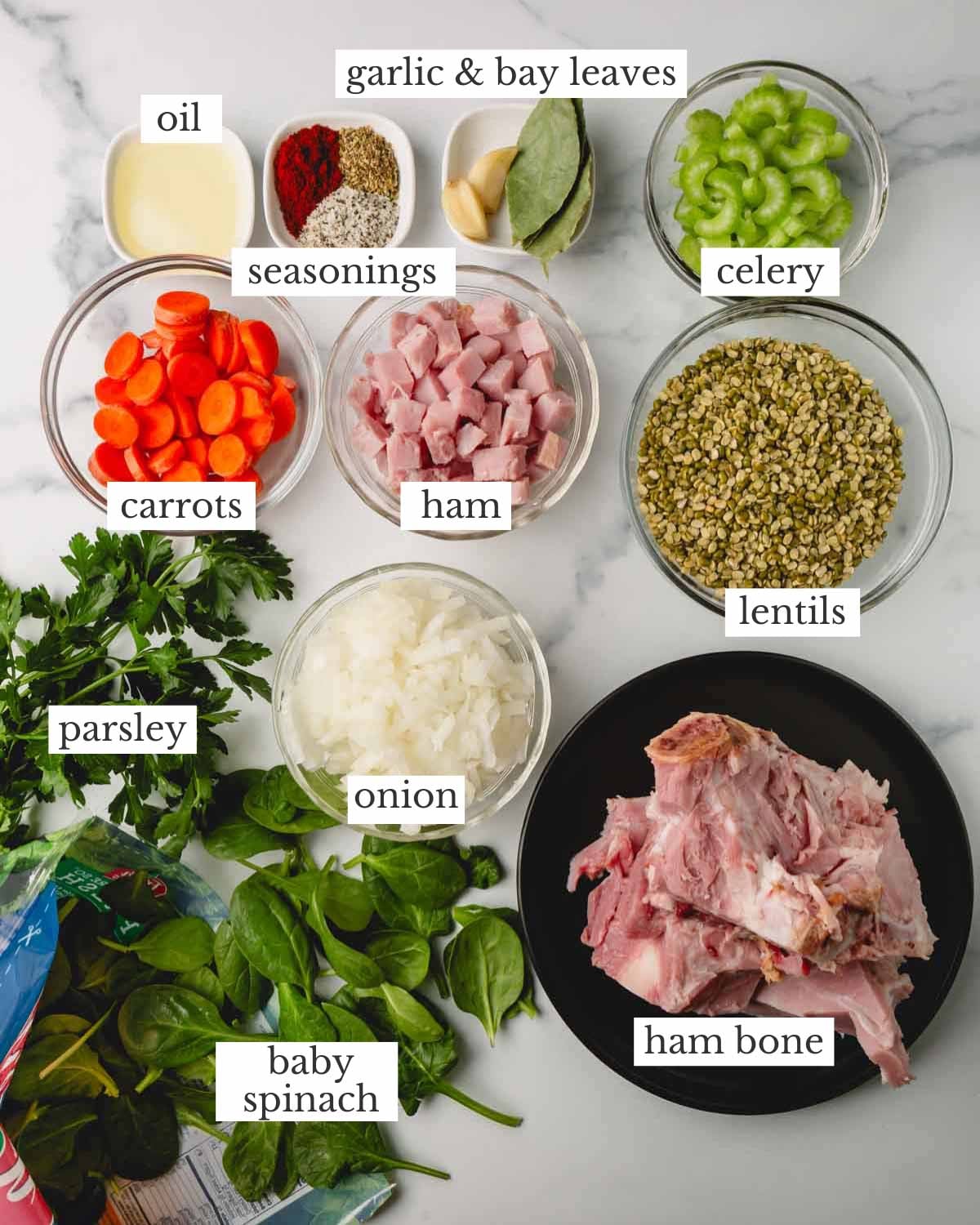 Ingredient shot for ham and lentil soup. The ingredients from top to bottom include, olive oil, seasonings, garlic cloves, bay leaves, celery, carrots, cubed ham, lentils, onion, parsley, baby spinach, and a ham bone.
