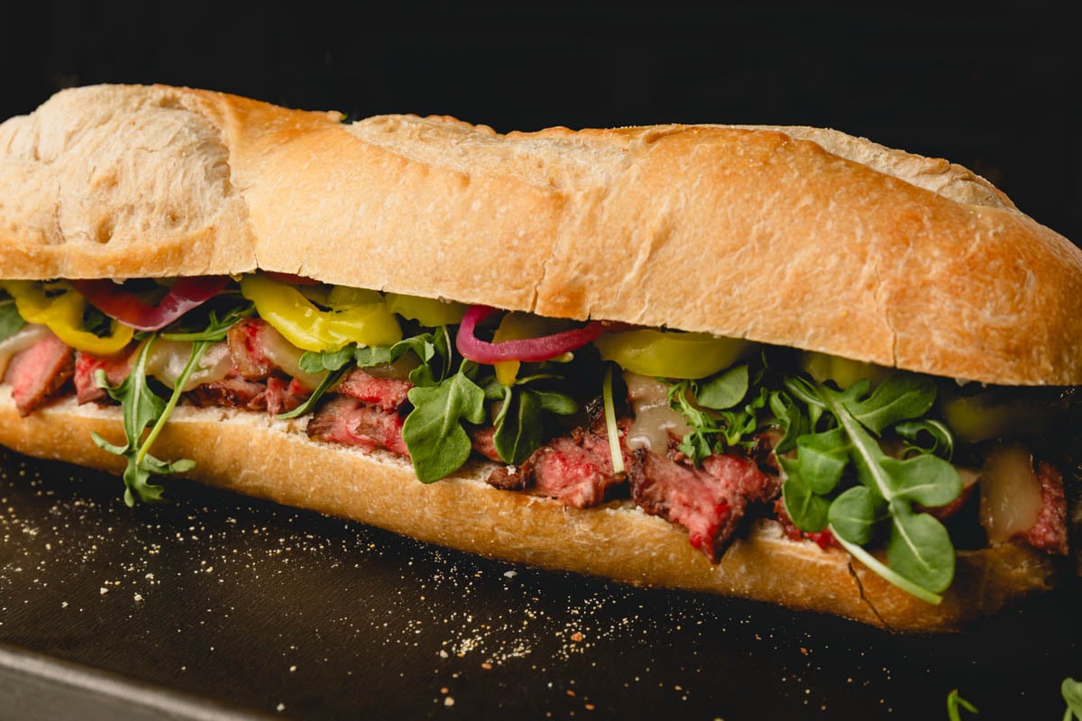 A cheese and ribeye steak sandwich with arugula, pickled red onion, and banana peppers on a baguette.