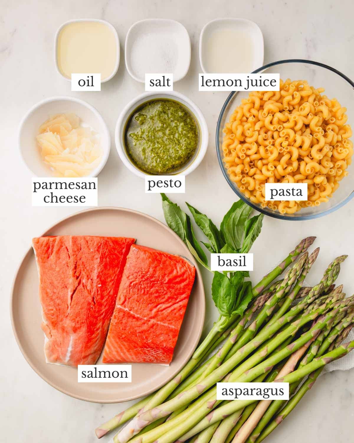 Ingredients for salmon pesto pasta. The bowls from top to bottom include olive oil, salt, lemon juice, parmesan cheese, pesto, pasta, salmon, basil, and asparagus.