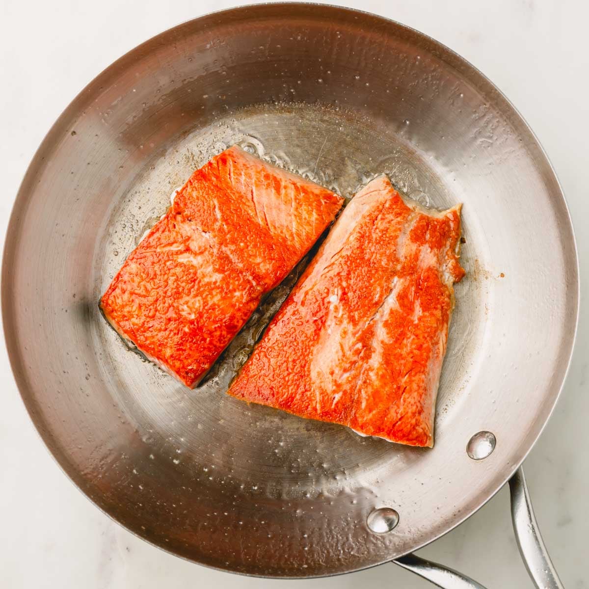 Two cooked salmon filets in a stainless steel skillet skin-side down.