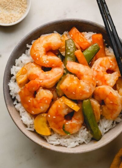 A close-up image of the finished shrimp teriyaki stir fry over white rice in a gray bowl with black chopsticks. A small white bowl with sesame seeds is in the back.