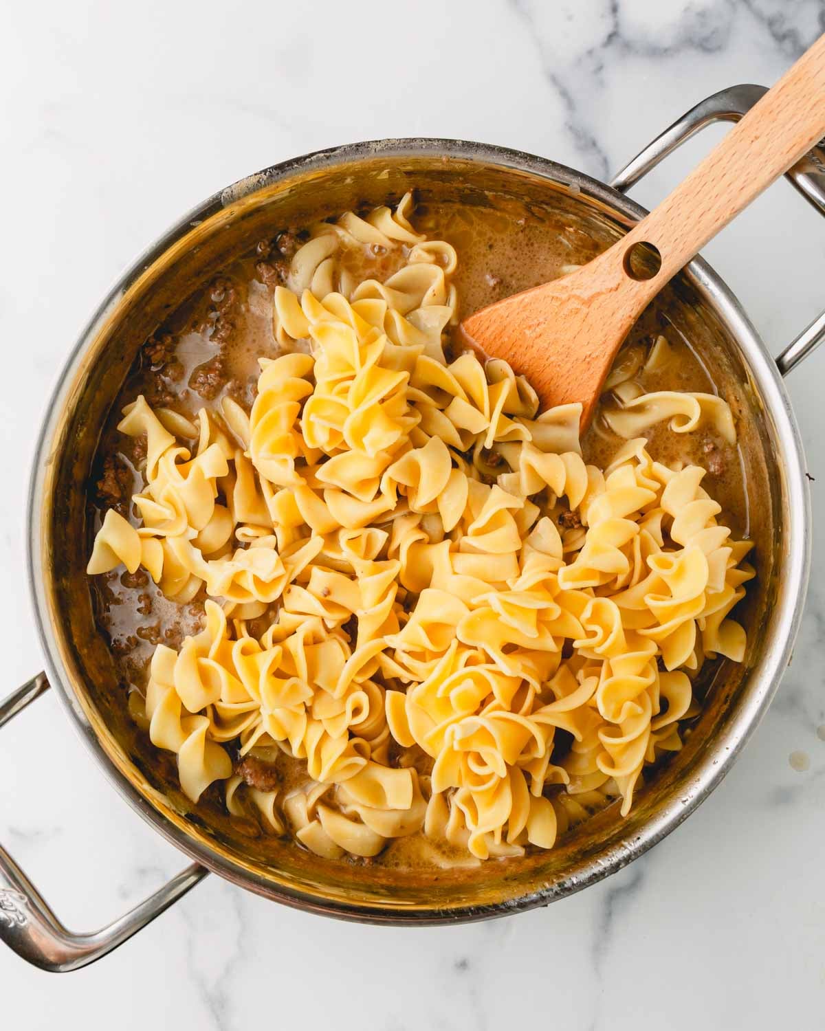 Cooked noodles are added to the sauce in a large skillet with a wooden spoon.