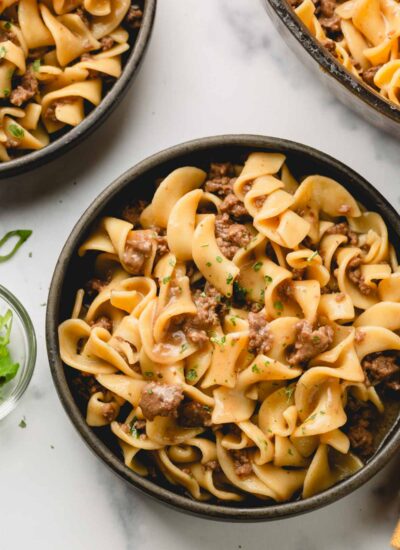 Ground beef and noodles are plated in a dark gray bowl. A second bowl as well as the skillet are in the background.