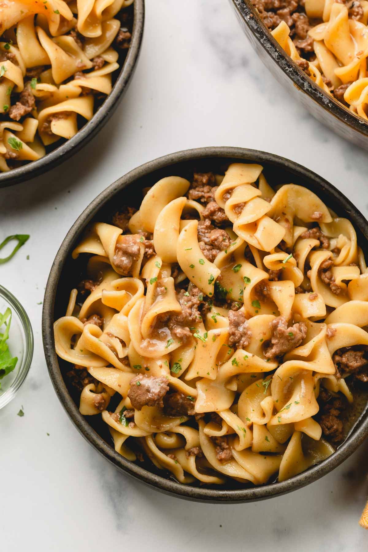 Ground beef and noodles are plated in a dark gray bowl. A second bowl as well as the skillet are in the background.