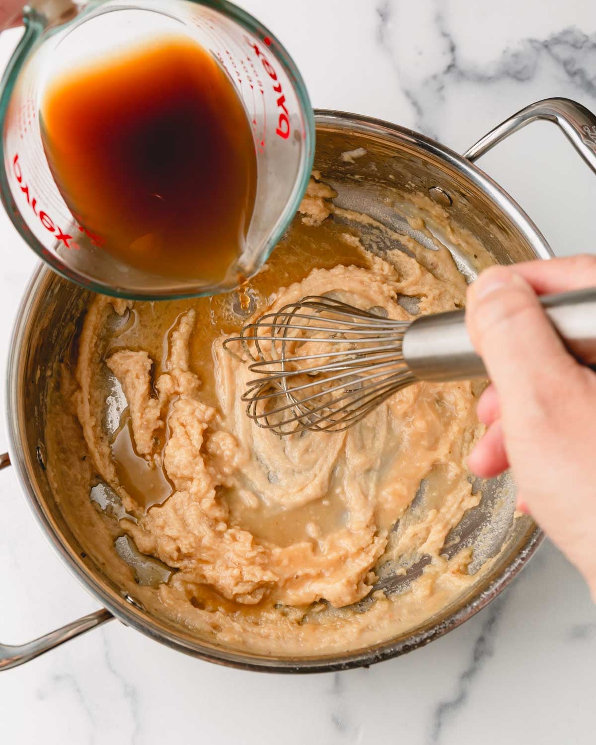 One hand is whisking with the other hand is adding beef broth from a glass measuring cup. The broth is being poured into the flour paste in the large skillet.