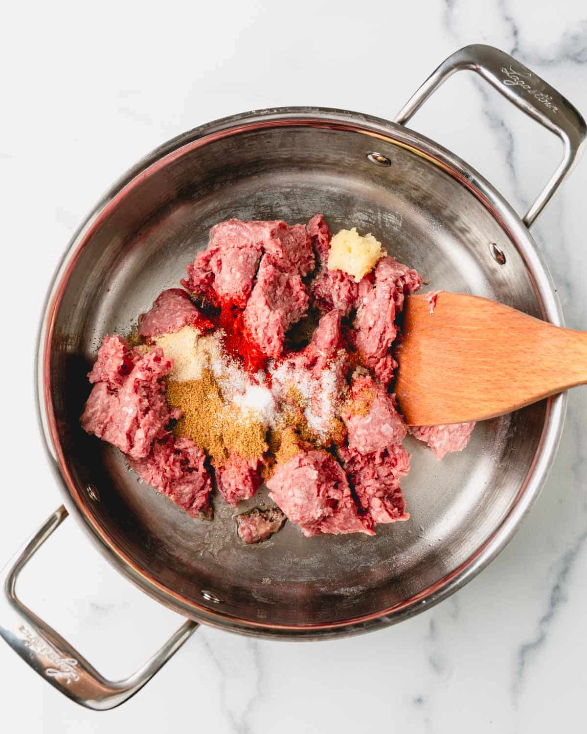 Ground beef is being cooked with smoked paprika, cumin, onion powder, garlic powder, salt, and garlic. A wooden spoon is in the beef.