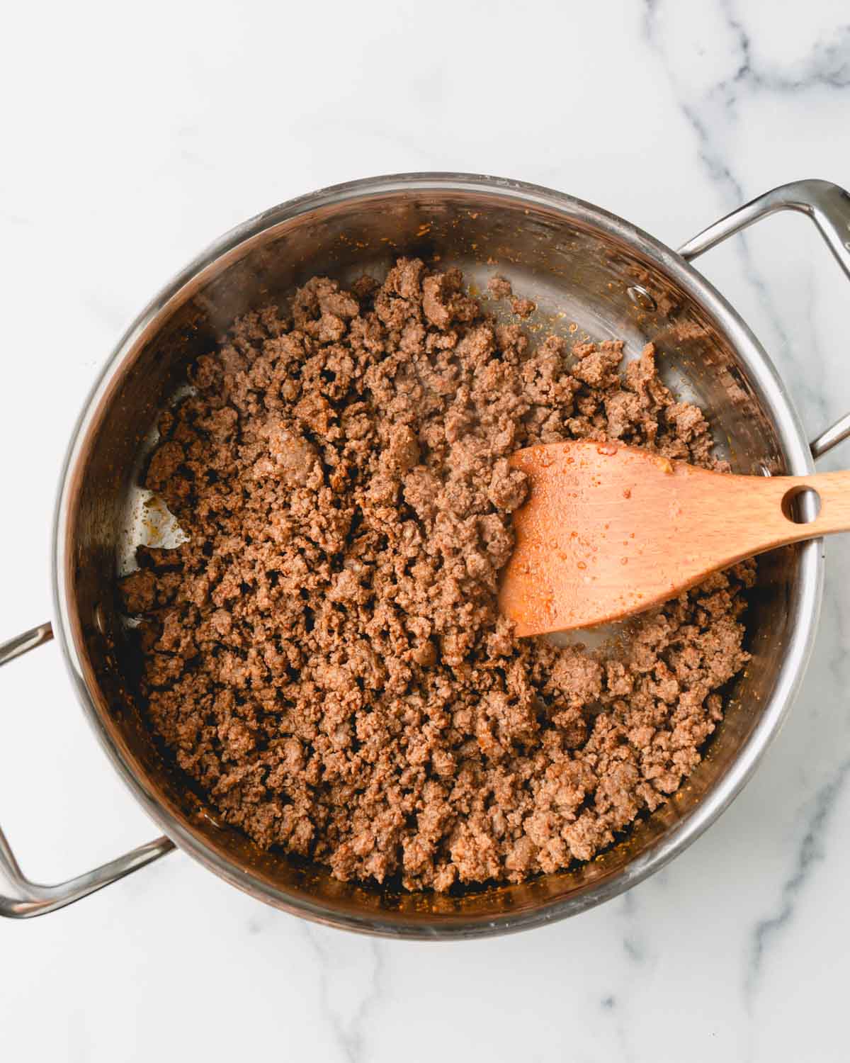 Ground beef is being browned in a large skillet using a wooden spoon.