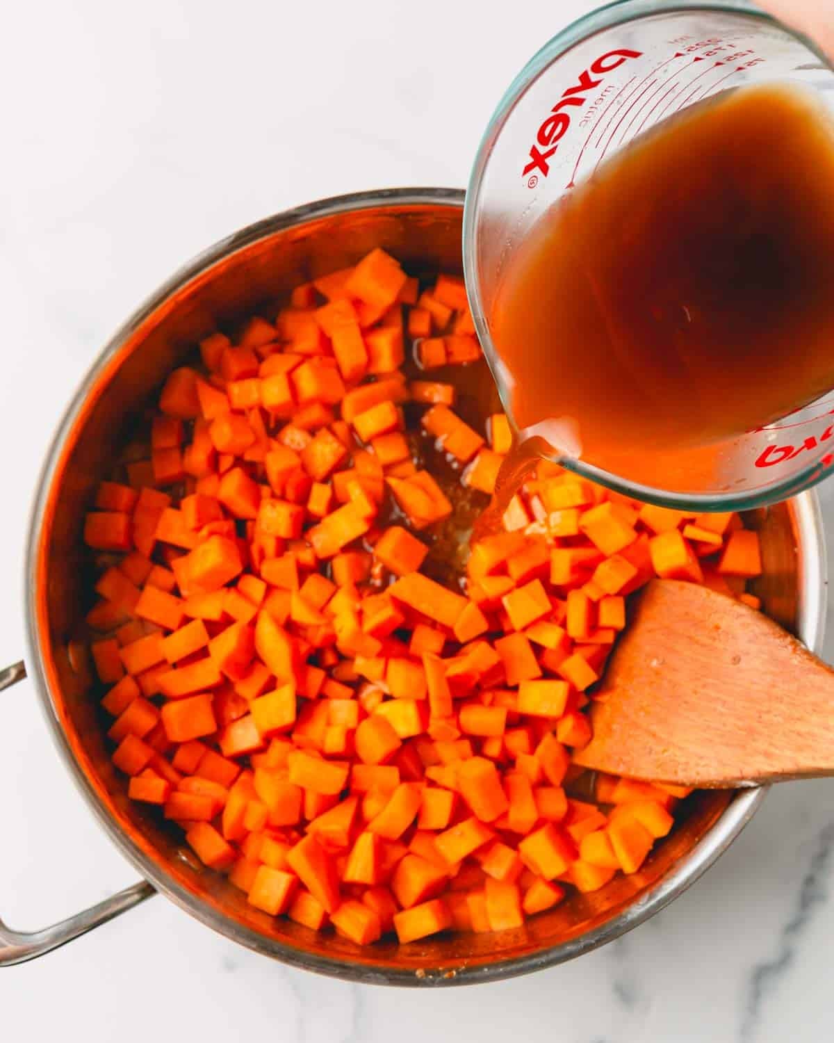 Beef broth is being added to sweet potatoes in a large skillet with a wooden spatula.