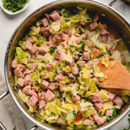 Ham and cabbage in a large saute pan with a wooden spoon. Parsley is in a white bowl in the background.
