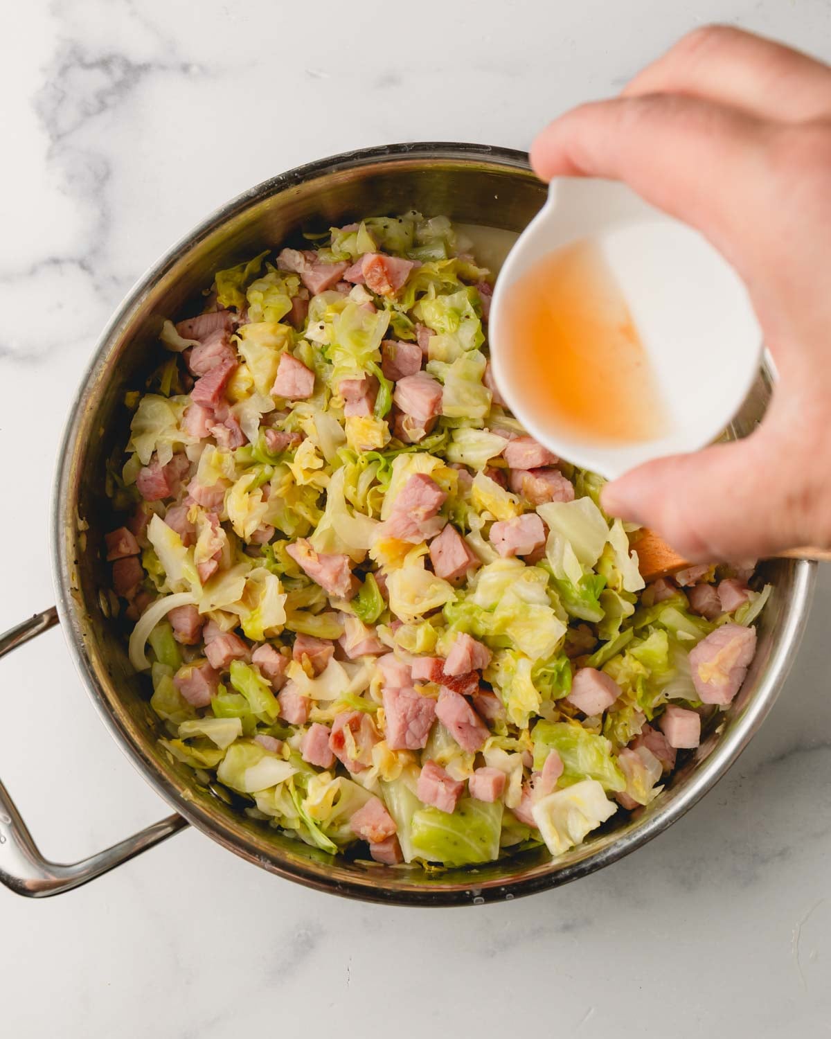 Vinegar is being poured into the saute pan by a hand. Cabbage and ham are cooking in the large saute pan.