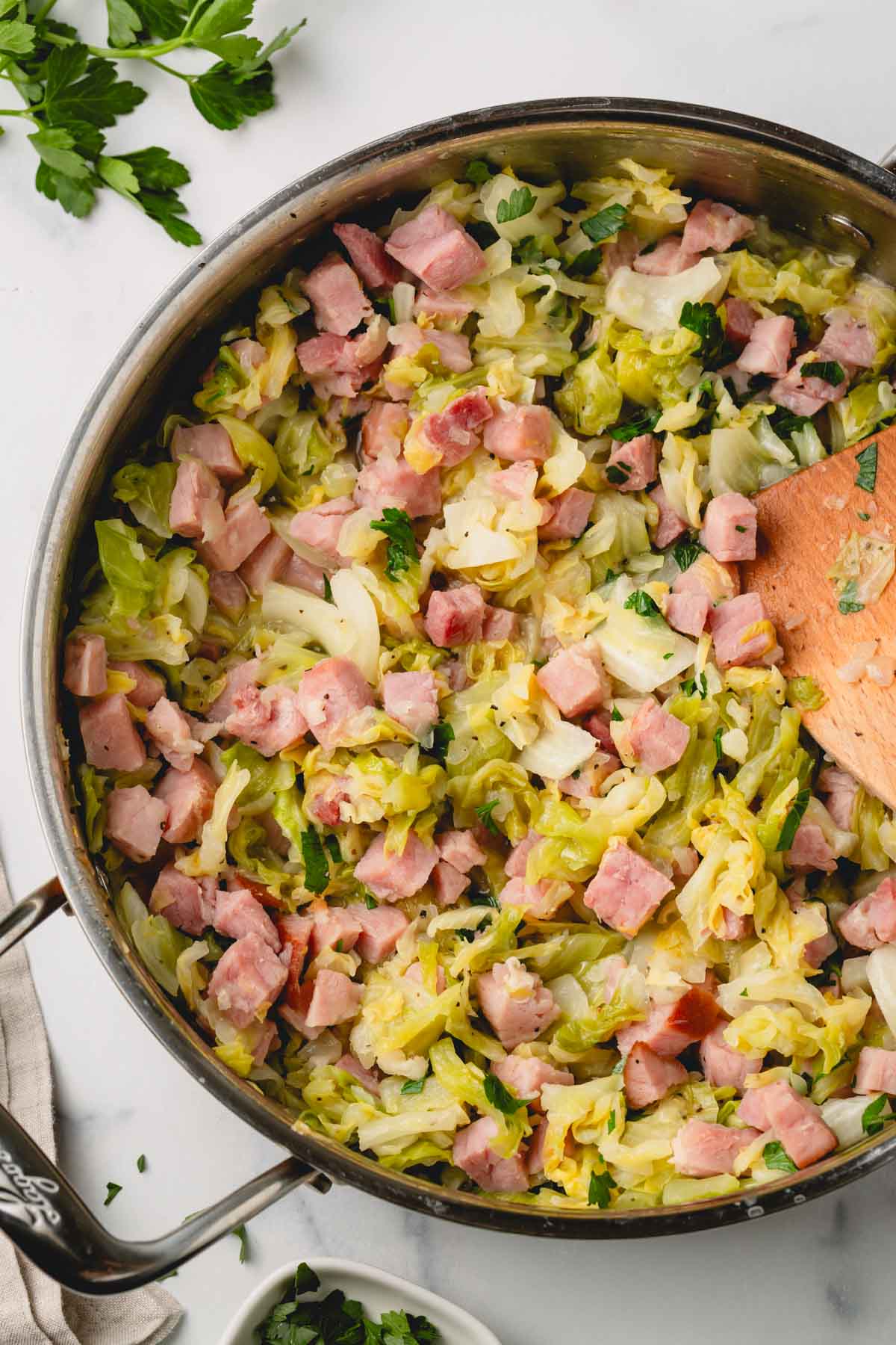 Parsley is added to the ham and cabbage that is cooking in a large saute pan with a wooden spoon.