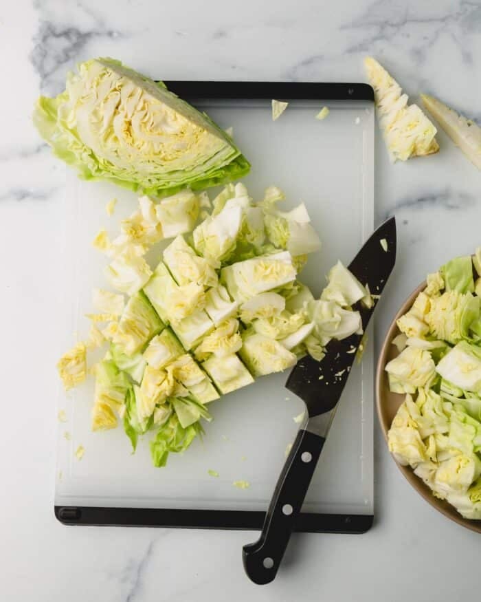 Cabbage is on a cutting board with a knife being chopped into pieces. Other pieces are in a brown bowl on the right.