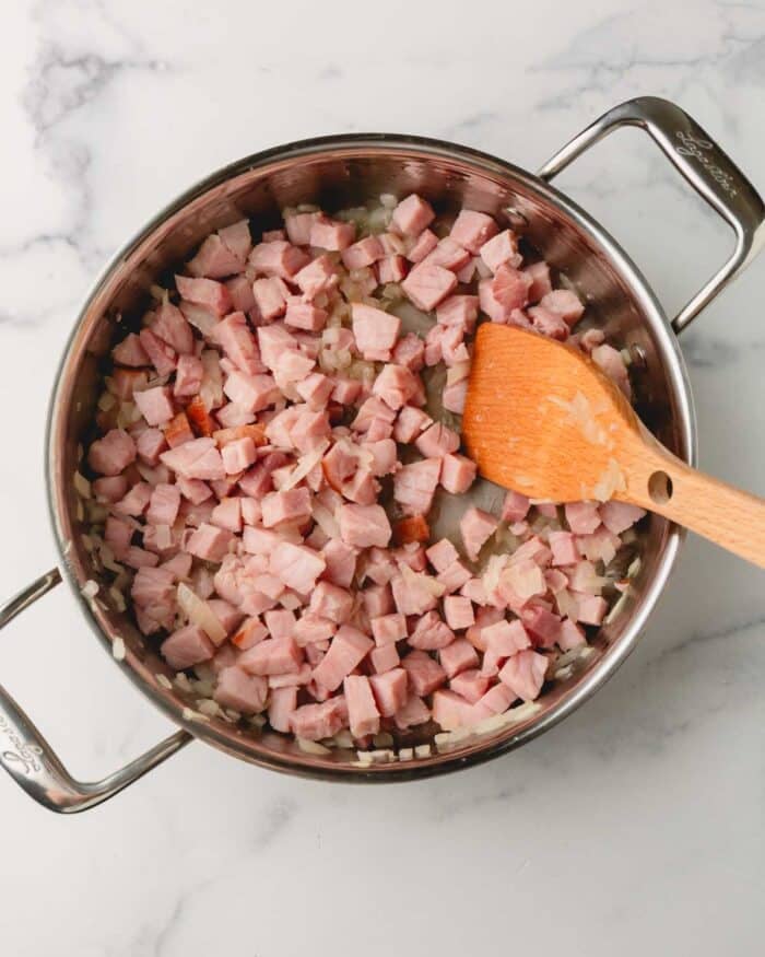 Ham and onion are cooking in a large saute pan with a wooden spoon.