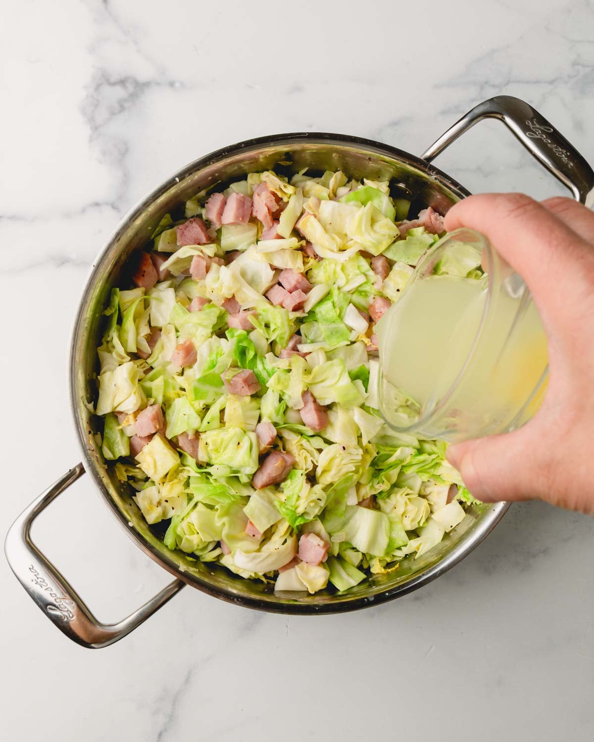 Chicken stock is being poured into the saute pan by a hand. Cabbage and ham are in the pan.