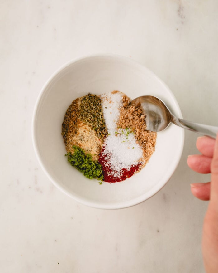 A hand with a spoon is mixing the blackening seasoning ingredients in a small bowl.