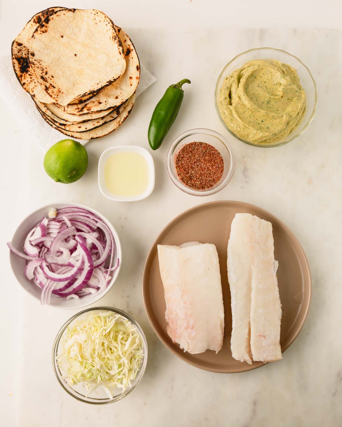 Ingredients for fish tacos which include corn tortillas, lime, jalapeno, grapeseed oil, blackening seasoning, avocado crema, red onion, green cabbage, and cod.