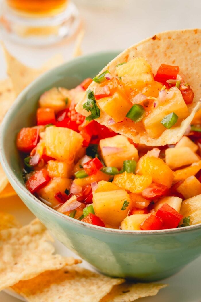A closeup image of a tortilla chip scooping up pineapple mango salsa from a serving bowl.