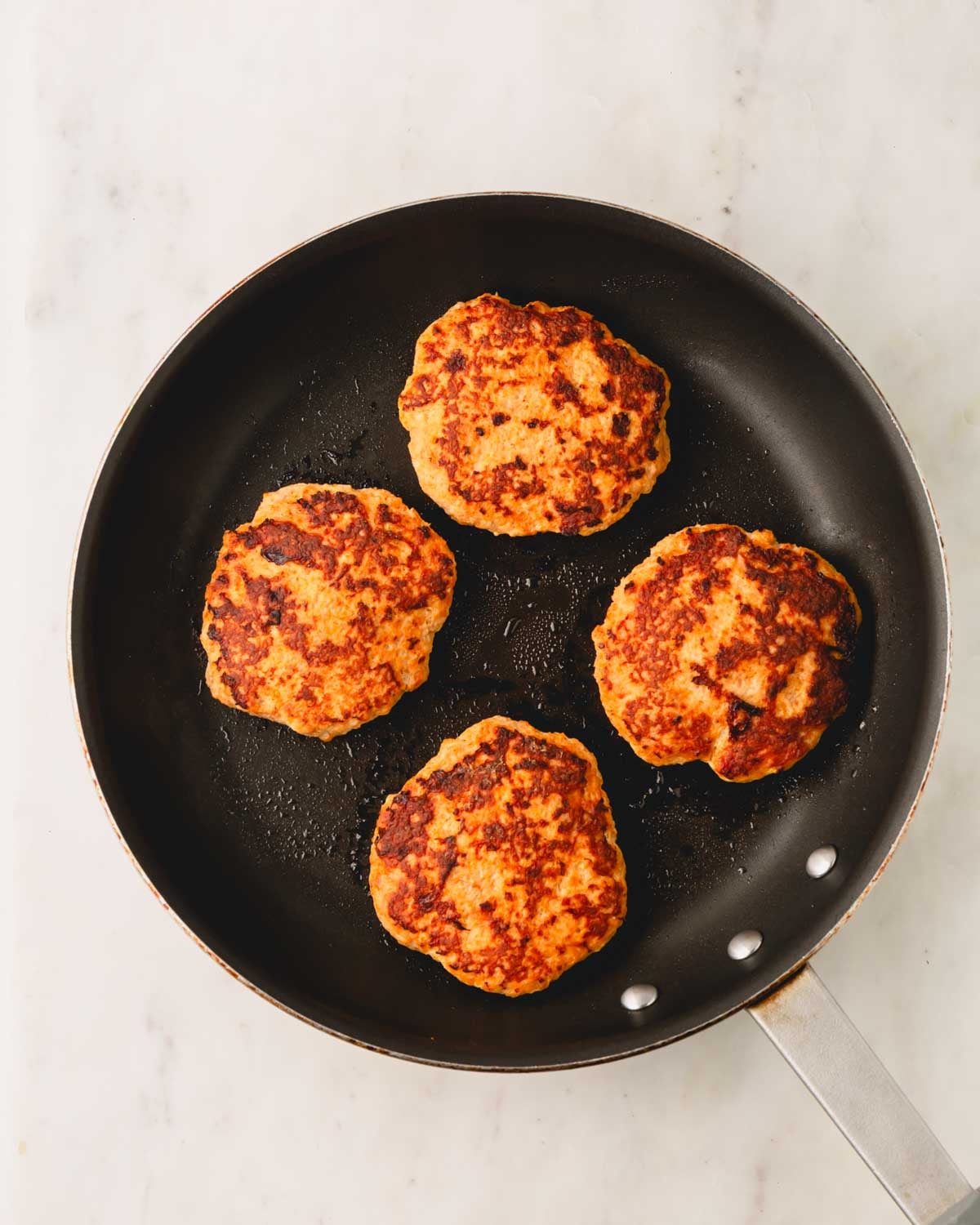 A large skillet has four spicy chicken burger patties cooking.