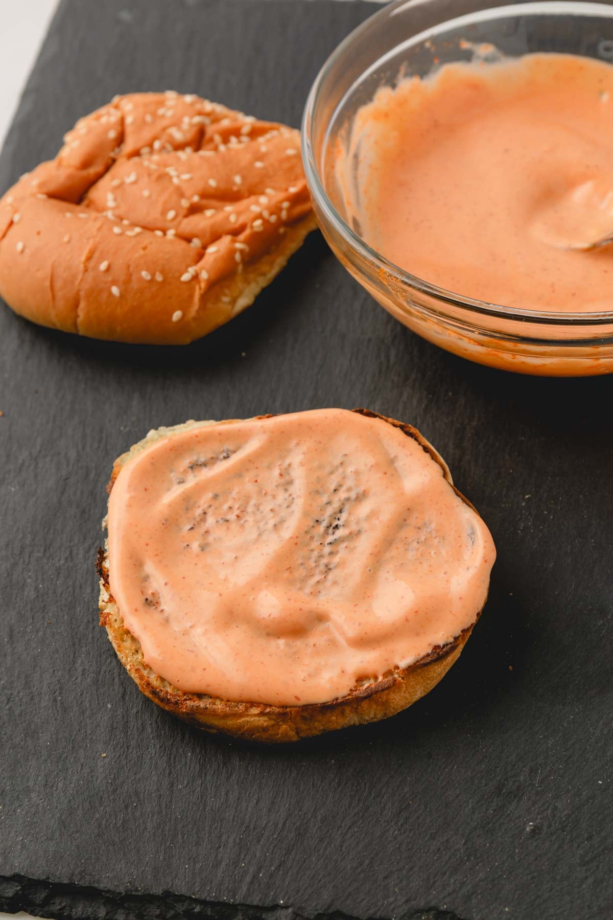 Spicy mayonnaise is spread onto a toasted burger bun. The bowl of spicy mayonnaise is in the background.